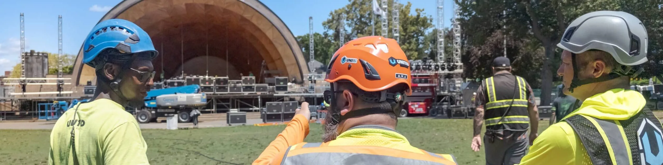 Three men with hi-vis shirts and hard hats stand on the grass pointing towards a stage