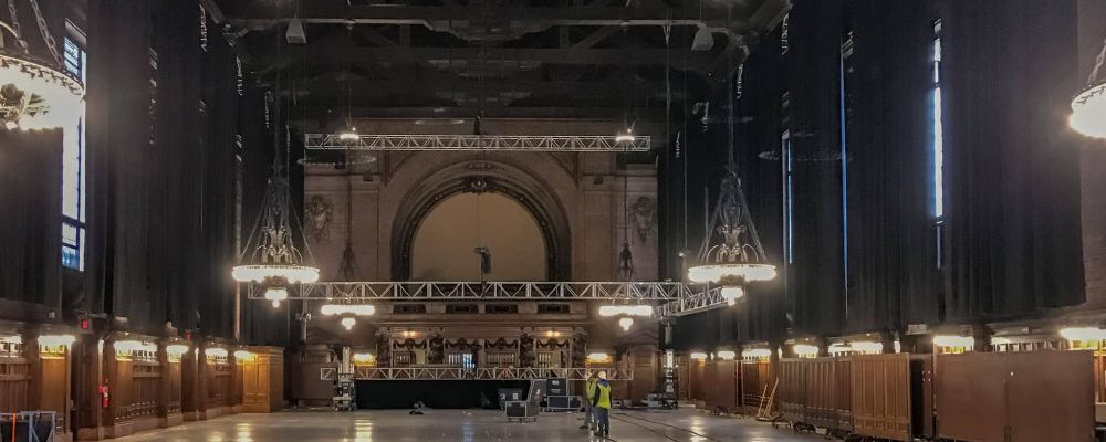 Black drape, which is attached to truss that lines the perimeter of teh cieling, hang down covering up the hall windows, blacking out the hall from natural light.