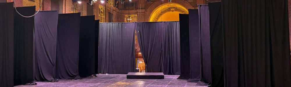 Inside a grand hall with the ceiling in a yellow wash and the floor in a pinkish-purple light wash. IN the middle are parallel rows of black drape on pipes going halfway up the height of teh room , against each wall, so the center is the open floor/stage.