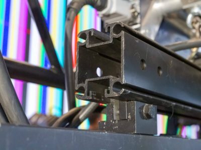 Upclose image of the end of a UNI-KEDER rail with a UNI-Clamp on the underside strut channel connecting the UNI-Keder to a video wall header (you cannot tell that the metal piece is a video wall header in this image though). Behind this is some more rigging connections and cropped metal pieces that are part of truss and a concert stage build. Behind everything there is a video wall showing a multi-colored, vertical striped test pattern. The focus though is on the UNI-Keder being used along with the uni-clamp.