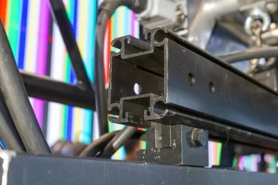 Upclose image of the end of a UNI-KEDER rail with a UNI-Clamp on the underside strut channel connecting the UNI-Keder to a video wall header (you cannot tell that the metal piece is a video wall header in this image though). Behind this is some more rigging connections and cropped metal pieces that are part of truss and a concert stage build. Behind everything there is a video wall showing a multi-colored, vertical striped test pattern. The focus though is on the UNI-Keder being used along with the uni-clamp.