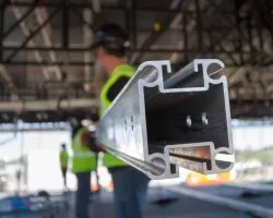 a couple unidentifiable stage workers in safety yellow vests hold up a piece of uni-keder while it gets attached to the underside of a stage roof