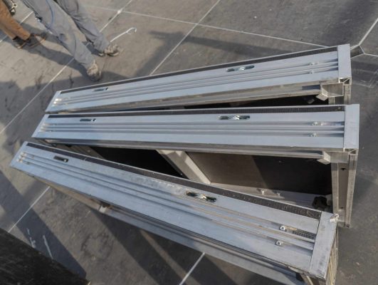 Looking down a the long side of 3 uni-dec which are vertical (on its long end) on an outdoor stage. The legs of a stage worker are in the upper left corner.