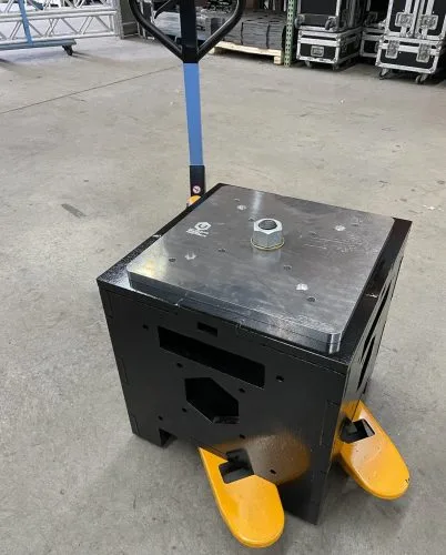 Uni-Bloc Jr, a black stage ballast patented by United Staging and Rigging, sits on top of a yellow and blue pallet jack to showcase the pallet jack pick points. POV is looking down at it so you can see the silver, adjustable top plate. United Staging & Rigging