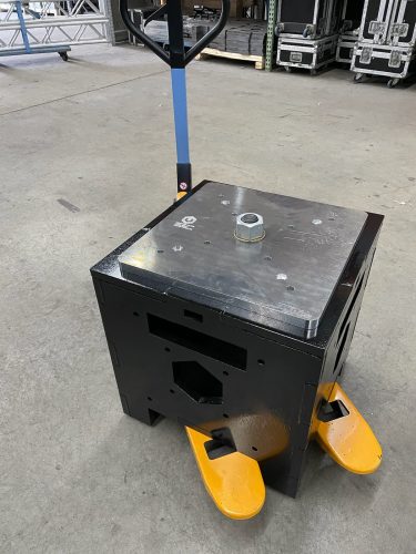Uni-Bloc Jr, a black stage ballast patented by United Staging and Rigging, sits on top of a yellow and blue pallet jack to showcase the pallet jack pick points. POV is looking down at it so you can see the silver, adjustable top plate. United Staging & Rigging