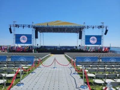 Umass Boston graduation stage, outdoors, on a sunny, cloudless day with a blue sky. The harbor ca be seen behind the stage in the middle right of the image. in front of the stage are rows of white, plastic, audience chairs and red ropes cordone off some of the plastic middle aisle walkway. The stage roof is yellow with a dark blue lighthouse and waves on it. A large video wall hangs down from truss on either side of the stage. The front of the stage is lined with flags from countries around the world.