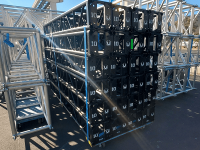 Stack of black 10 foot, 12 inch truss, surrounded by stacks of silver 20x30 inch trusses. outdoors. sunny day. black truss has "10" and the United Staging & Rigging "U"spray painted on the end of each stick of truss.