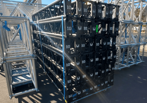 Stack of black 10 foot, 12 inch truss, surrounded by stacks of silver 20x30 inch trusses. outdoors. sunny day. black truss has "10" and the United Staging & Rigging "U"spray painted on the end of each stick of truss.