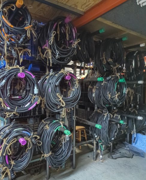 A wall of neatly coiled black motor cables for rigging chain hoists, hang in a warehouse. They are organized in color-codes by size.