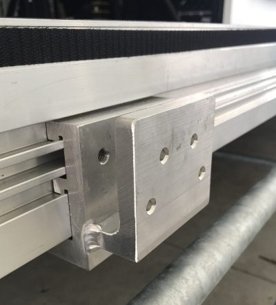 Metal flat cupping accessory piece hooked into the custom extrusion on the outside of United Staging & Rigging's patented Uni-Dec stage decking. This piece has four small holes in the front for attaching things. It is silver in to match the sides of the Uni-Dec